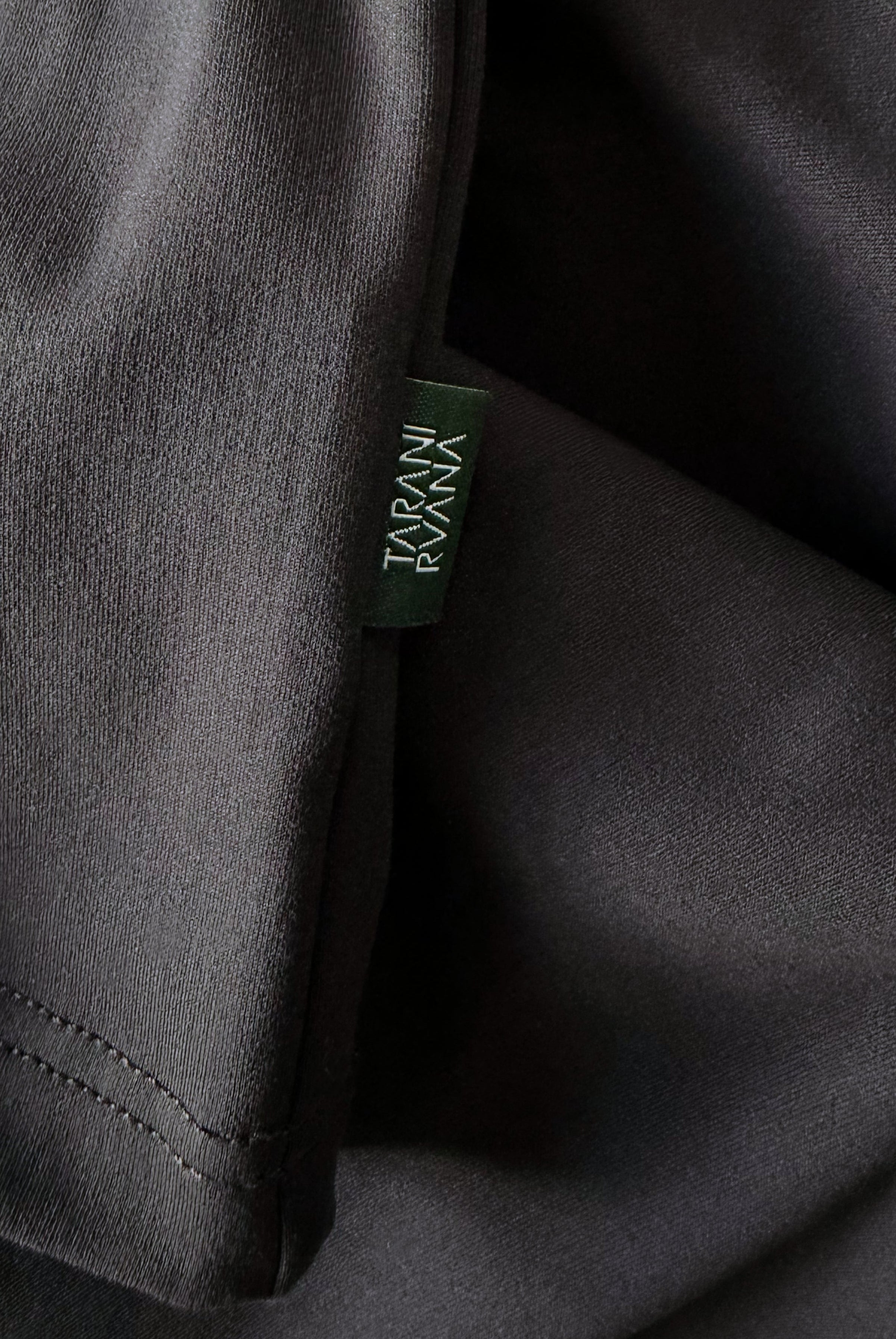Close-up view of TARANIRVANA Aria Polo shirt hem in Phantom black, highlighting the premium stitching and signature green brand woven label with quick-dry, moisture-wicking, odor-resistant, and UPF 50+ sun protection.