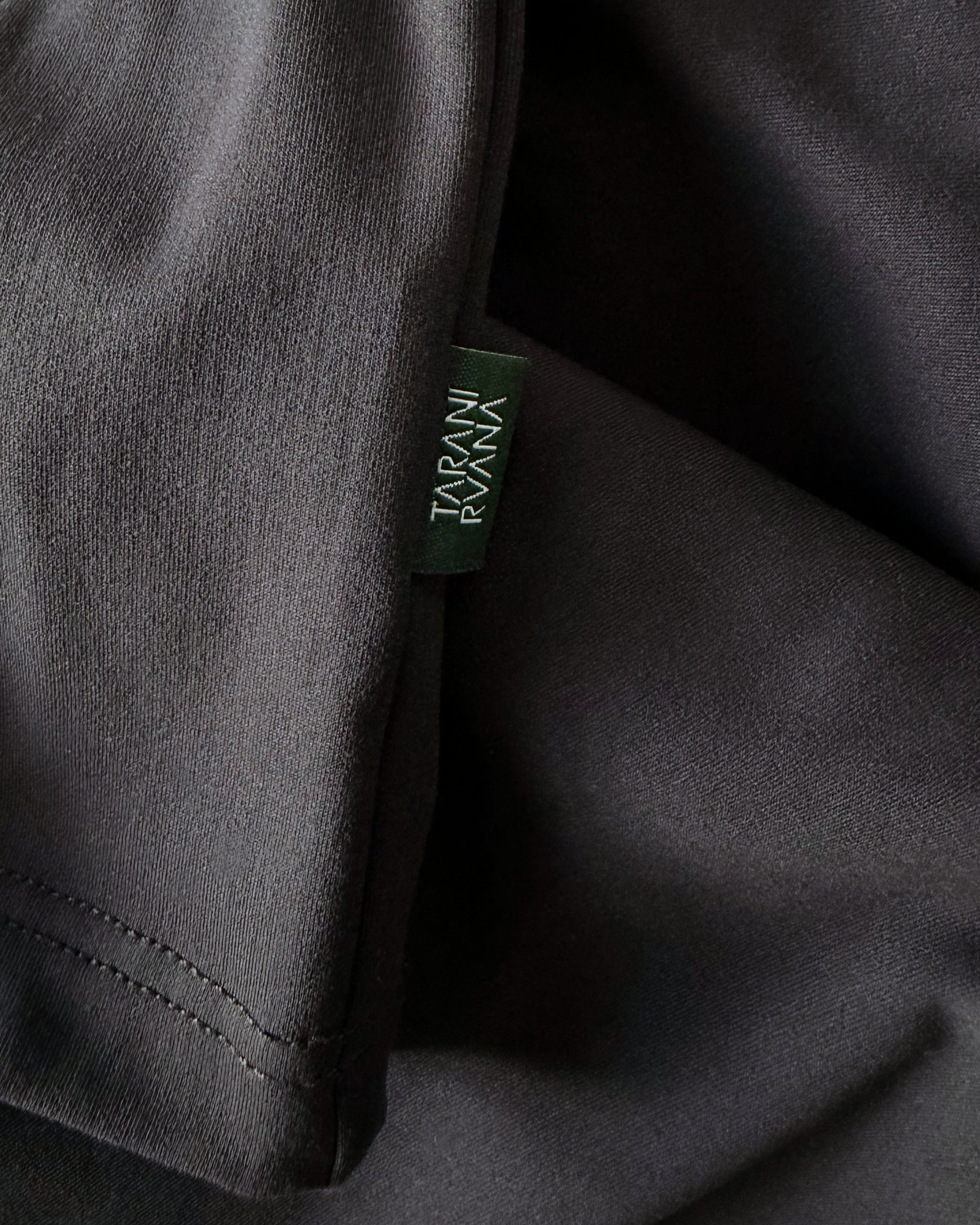 Close-up view of TARANIRVANA Aria Polo shirt hem in Phantom black, highlighting the premium stitching and signature green brand woven label with quick-dry, moisture-wicking, odor-resistant, and UPF 50+ sun protection.