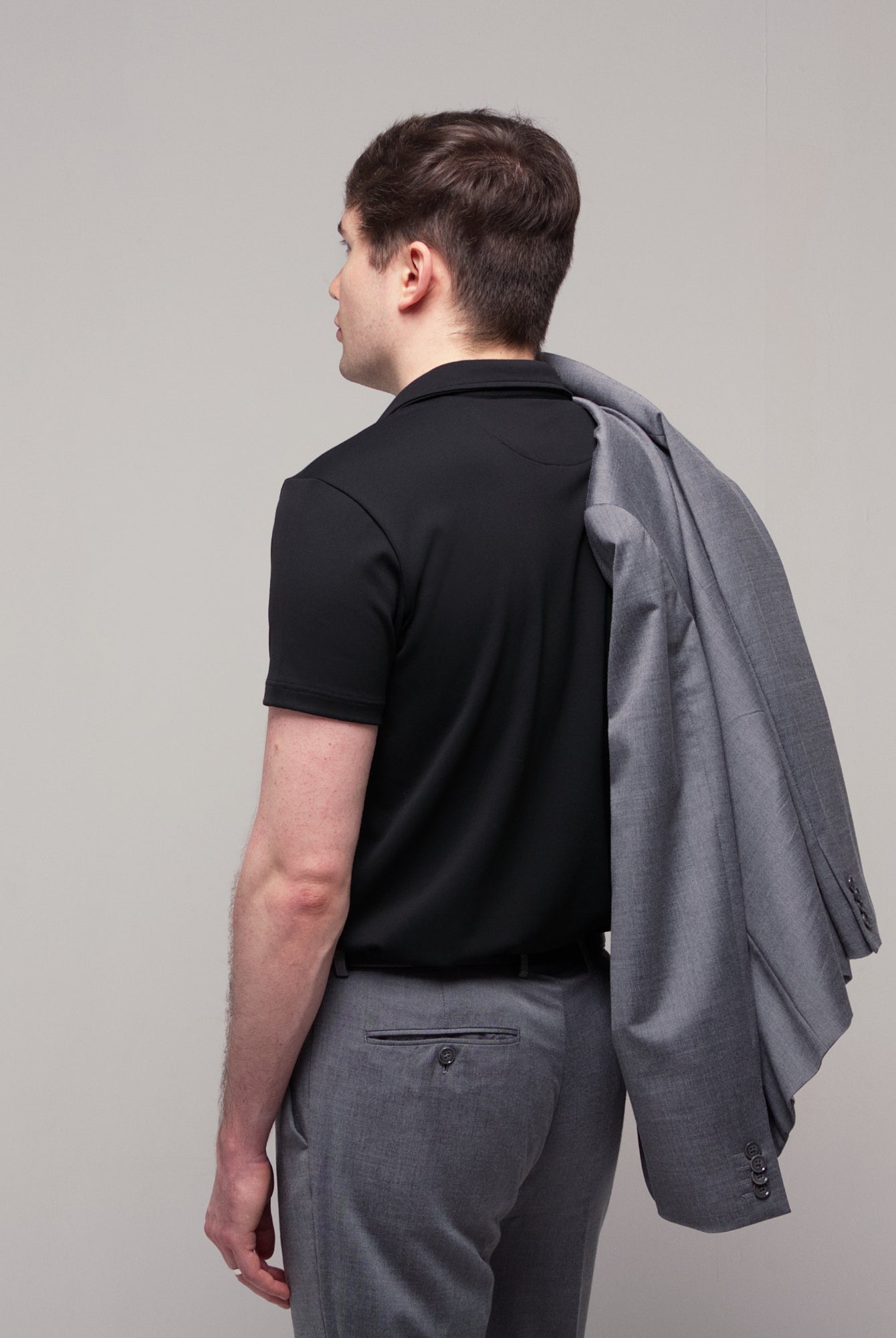 Back and side view of a man wearing TARANIRVANA Aria Polo in Phantom black with a gray suit jacket over his shoulder, showcasing high-performance SpiroTex™ fabric with quick-dry, moisture-wicking, odor-resistant, and UPF 50+ sun protection.