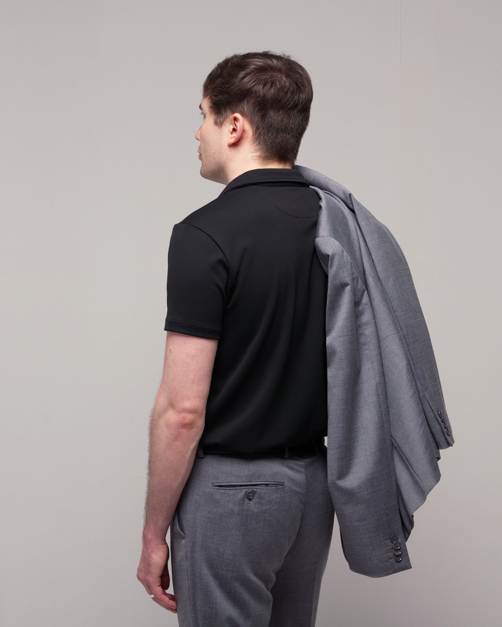 Back and side view of a man wearing TARANIRVANA Aria Polo in Phantom black with a gray suit jacket over his shoulder, showcasing high-performance SpiroTex™ fabric with quick-dry, moisture-wicking, odor-resistant, and UPF 50+ sun protection.