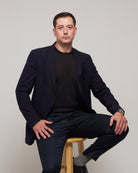Front view of a man styling TARANIRVANA Essenza T-Shirt in Phantom black layered under a sharp navy business suit jacket, featuring premium SpiroTex fabric with quick-dry, moisture-wicking, odor-resistant, and UPF 50+ sun protection.